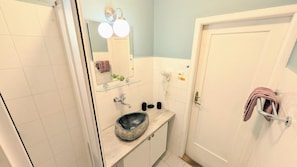A bathroom with a stone sink, a glass shelf, a mirror with lights, a towel rack, and a door.