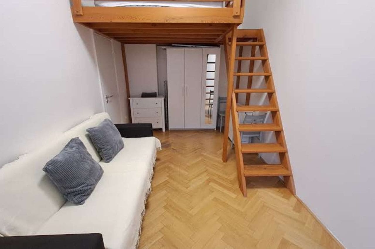 A small room with a bunk bed, a wooden ladder, a white sofa with two pillows, and a herringbone-patterned wooden floor.