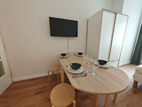 A dining area with a long wooden table set for six, a flat-screen TV mounted on the wall, and a white wardrobe in the background.
