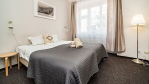 A hotel room with a bed, a small table, a lamp, a window with curtains, and a framed picture on the wall.