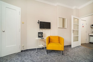 A room with a yellow armchair, a flat-screen TV mounted on the wall, a small table with a speaker, and a door leading to another room. - Cozy studio with terrace by tram (Praha 3)
