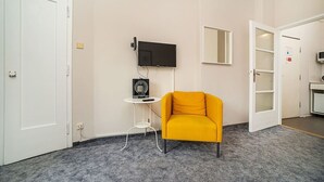 Quarto com poltrona amarela, TV de tela plana na parede, mesa pequena com um alto-falante e porta que dá para outro cômodo.