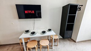A dining area with a white table, four wooden chairs, a black TV mounted on the wall displaying 'NETFLIX', and a black cabinet to the right.