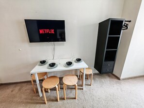 A dining area with a white table, four wooden chairs, a black TV mounted on the wall displaying 'NETFLIX', and a black cabinet to the right.