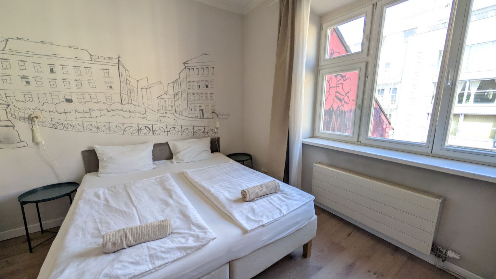 Un lit double avec du linge de lit blanc, un radiateur, une fenêtre avec des rideaux et un mur orné d’une esquisse représentant un paysage urbain.