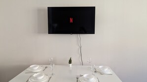 A dining table set for four with white plates, glasses, and cutlery, a small potted plant, and a flat-screen TV mounted on the wall displaying the Netflix logo.