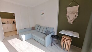 A modern living room with a light blue sectional sofa, a small black table, and wooden chairs. A macrame wall hanging is visible on the green wall.