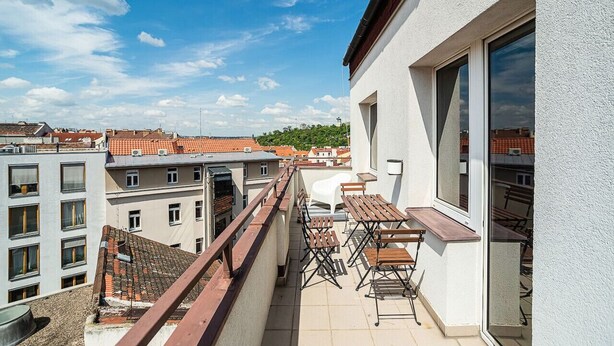 A balcony with outdoor furniture, a view of neighboring buildings, and a clear sky.