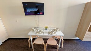 A dining area with a white table set for four, a television mounted on the wall displaying a Netflix sign, and a small potted plant on the table.