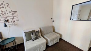 A small, neatly arranged room with two white upholstered chairs, a teal chair, a wall-mounted lamp, and a mirror reflecting a building.