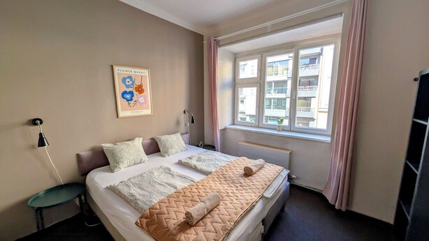 A hotel room with a bed, a nightstand, a window with curtains, and a wall with a framed picture.