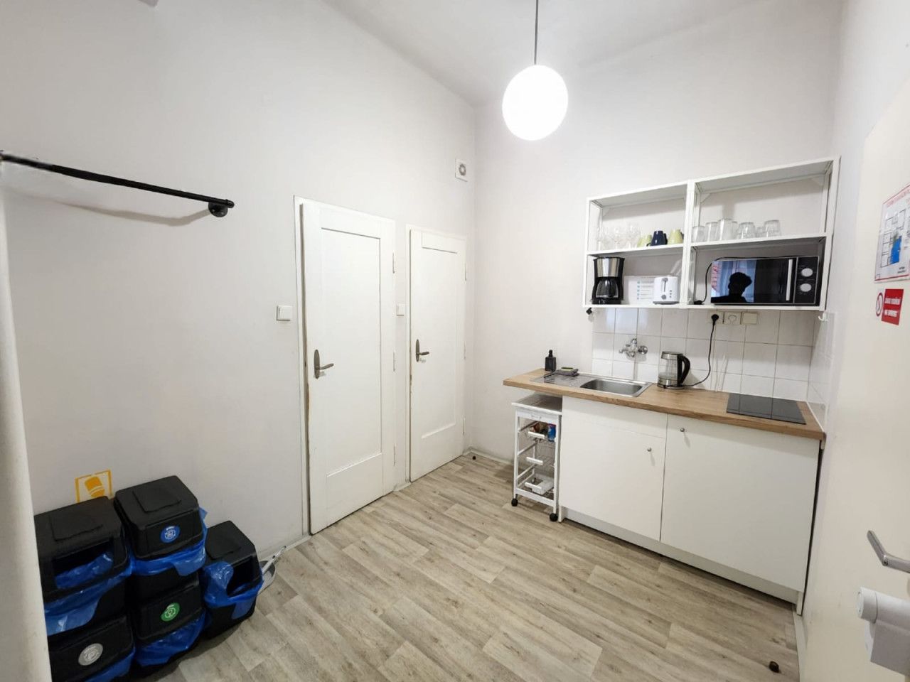 A compact kitchen with white cabinets, a wooden countertop, a microwave, a coffee maker, and a small sink. There are two white doors and a ceiling light.