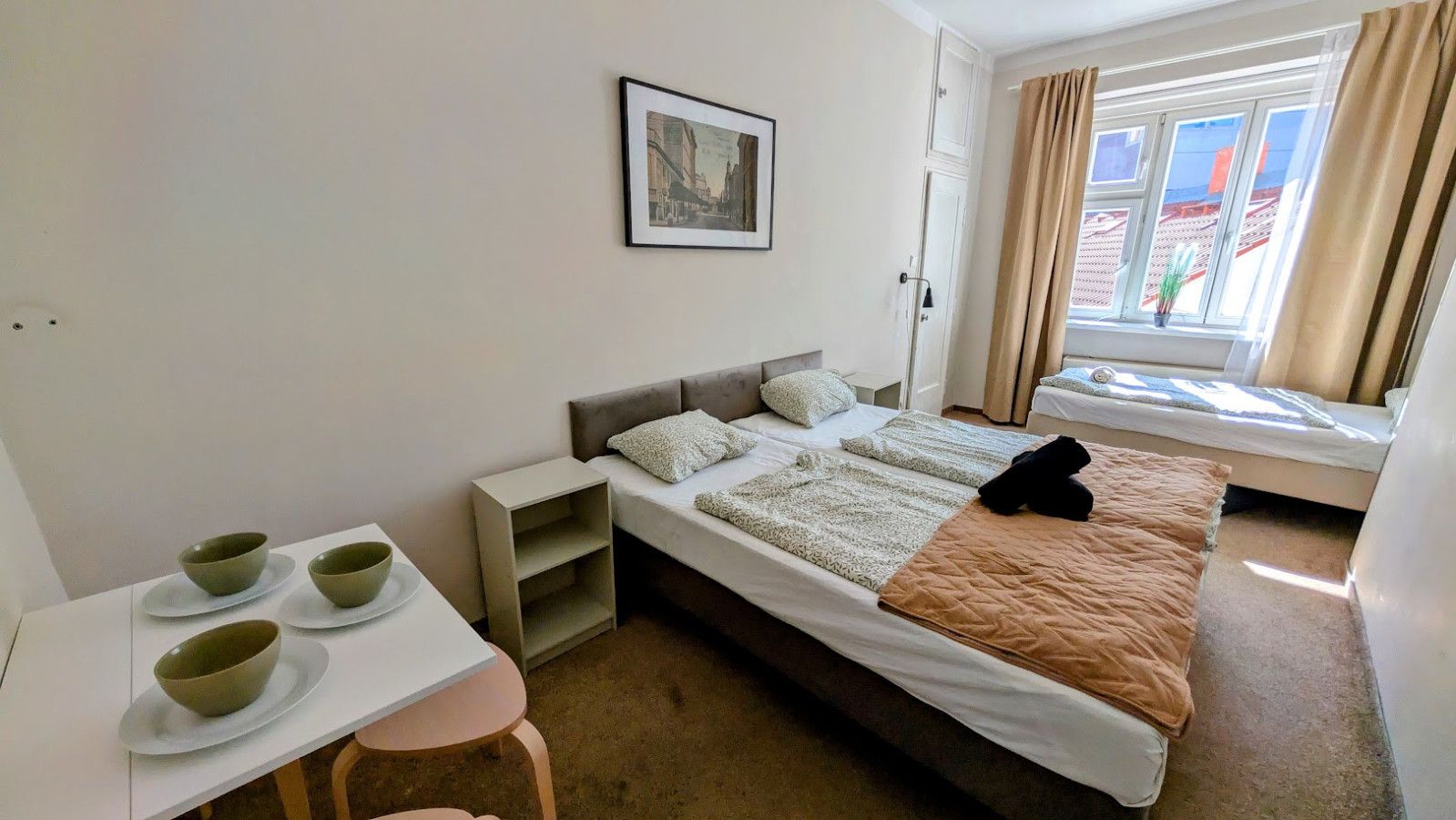 A hotel room with a bed, a desk with bowls, a chair, and a window with curtains.