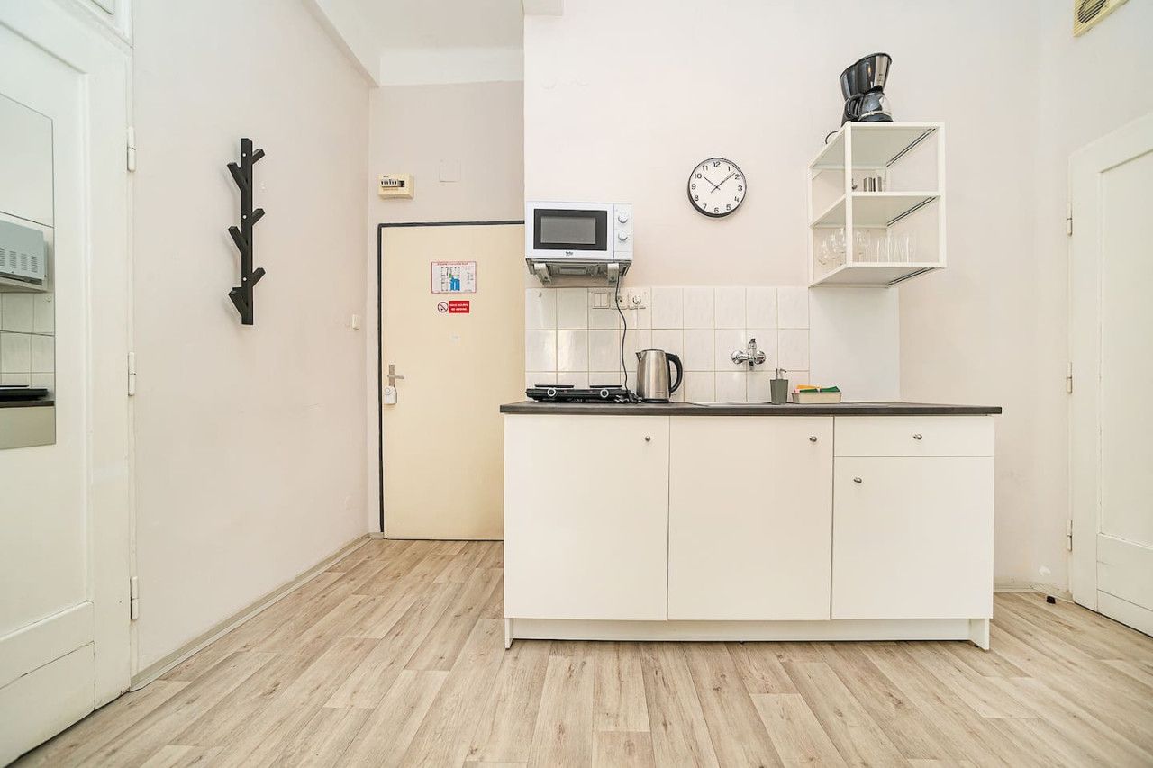 A compact kitchen with white cabinets, a microwave, a kettle, and a clock on the wall.