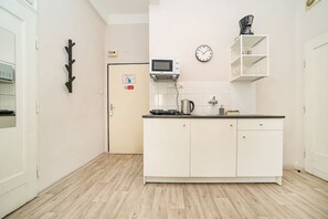A compact kitchen with white cabinets, a microwave, a kettle, and a clock on the wall.