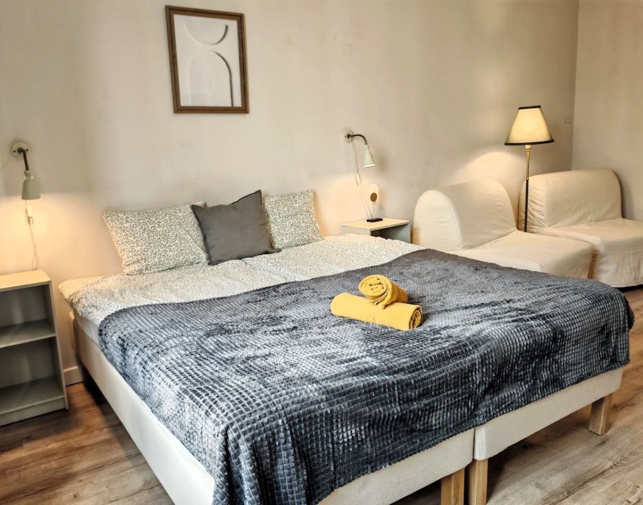 A neatly made bed with a patterned blanket and a rolled towel, a bedside table with a lamp, and a chair with a lamp.
