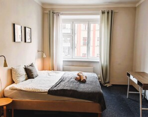 A bedroom with a bed, a bedside table, a lamp, and a window with curtains. - Clean apartment easy location (Praha 3)