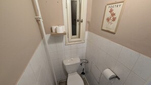 A small, narrow bathroom with a toilet, a sink, and a window. There is a shelf with toilet paper and a framed picture on the wall. - Modern apartment Great location for 7 people (Praha)