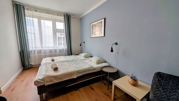 Une chambre à coucher avec un lit, une petite table ronde, une chaise, une lampe et une fenêtre avec des rideaux.