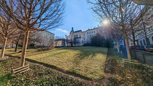 A grassy park area with benches and leaf-covered ground, surrounded by buildings with classical architecture.