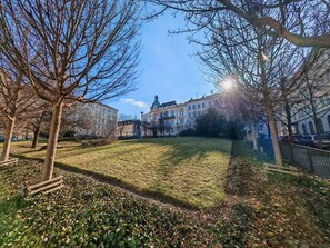 A grassy park area with benches and leaf-covered ground, surrounded by buildings with classical architecture.
