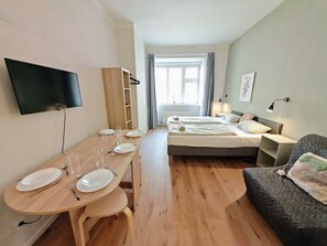 A hotel room with a wooden dining table set for two, a bed with a grey quilt, a TV mounted on the wall, and a window with curtains.