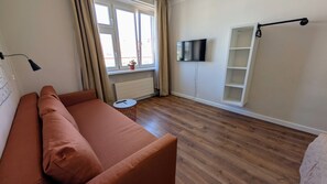A room with a wooden floor, a sofa, a TV mounted on the wall, and a window with curtains.