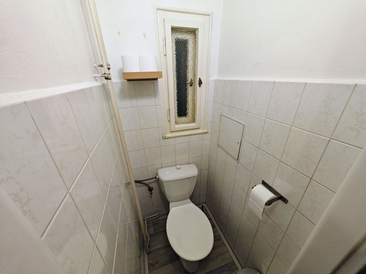 A small, narrow bathroom with a toilet, a window, and a towel rack.