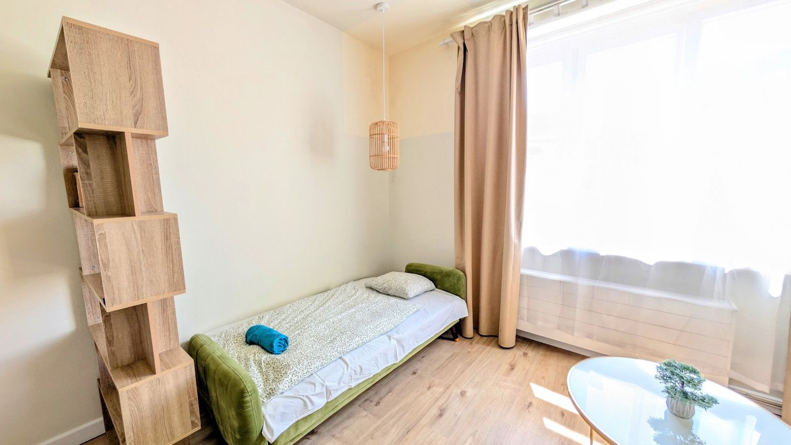 Ein kleines Zimmer mit einem Einzelbett, einer Holzregaleinheit, einer Pendelleuchte und einem Fenster mit Vorhängen.