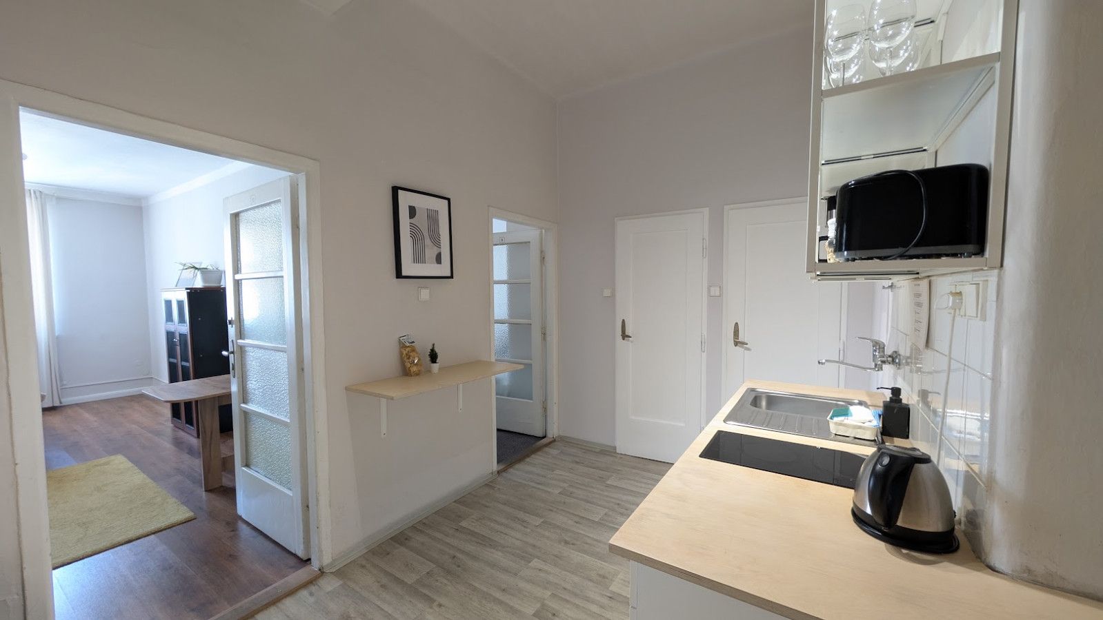 A modern kitchen with a wooden countertop, a kettle, and a microwave. There is a door leading to a hallway with a bench and a framed picture on the wall.