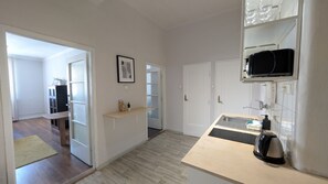 A modern kitchen with a wooden countertop, a kettle, and a microwave. There is a door leading to a hallway with a bench and a framed picture on the wall.