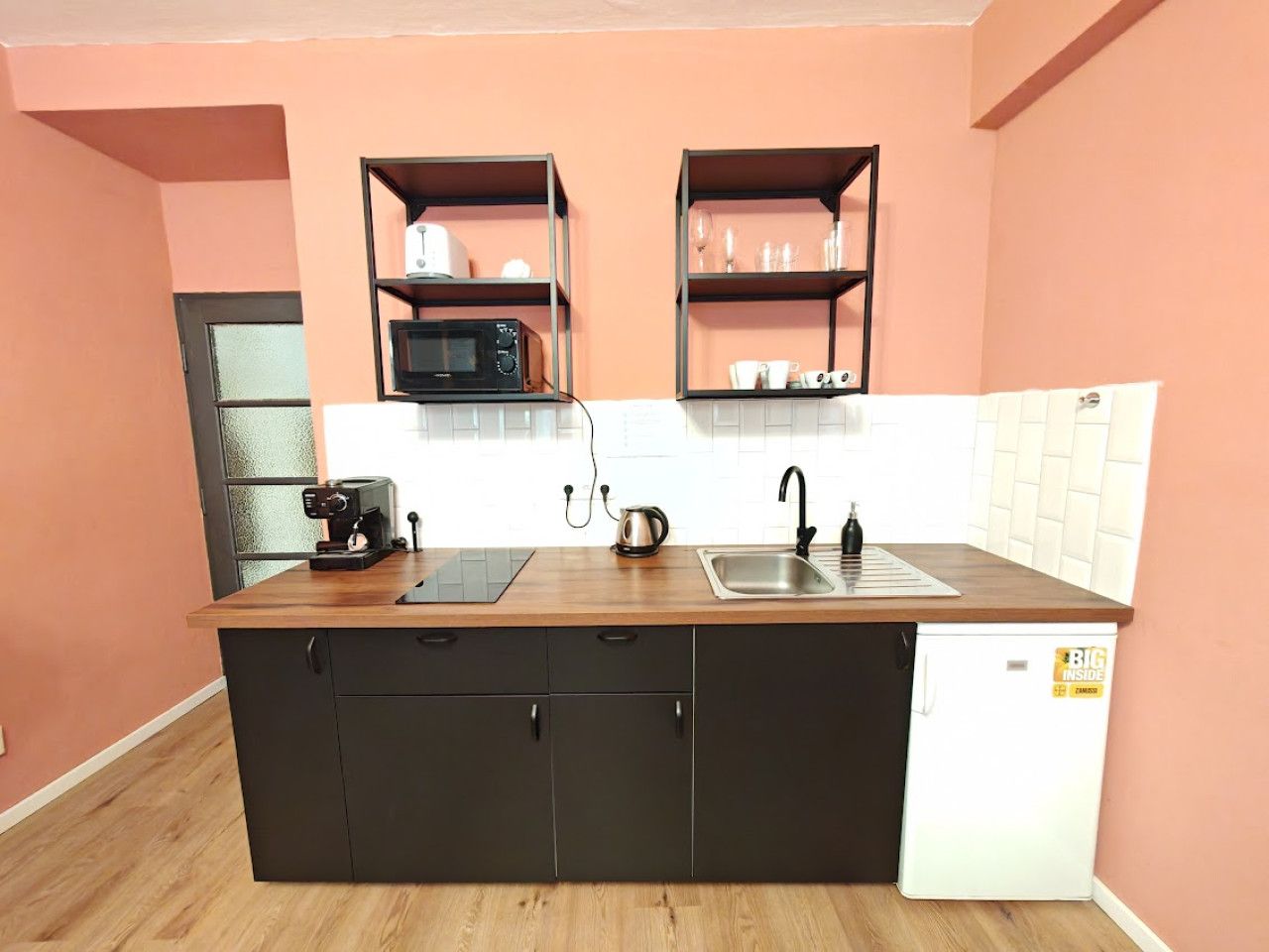 Cozinha compacta com bancada de madeira, armários pretos, micro-ondas, chaleira e pia.