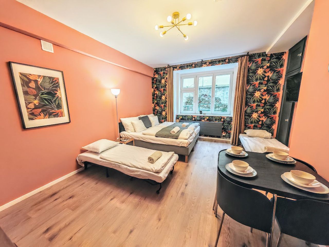 A room with two beds, a dining table with bowls, a painting, and a window with curtains.