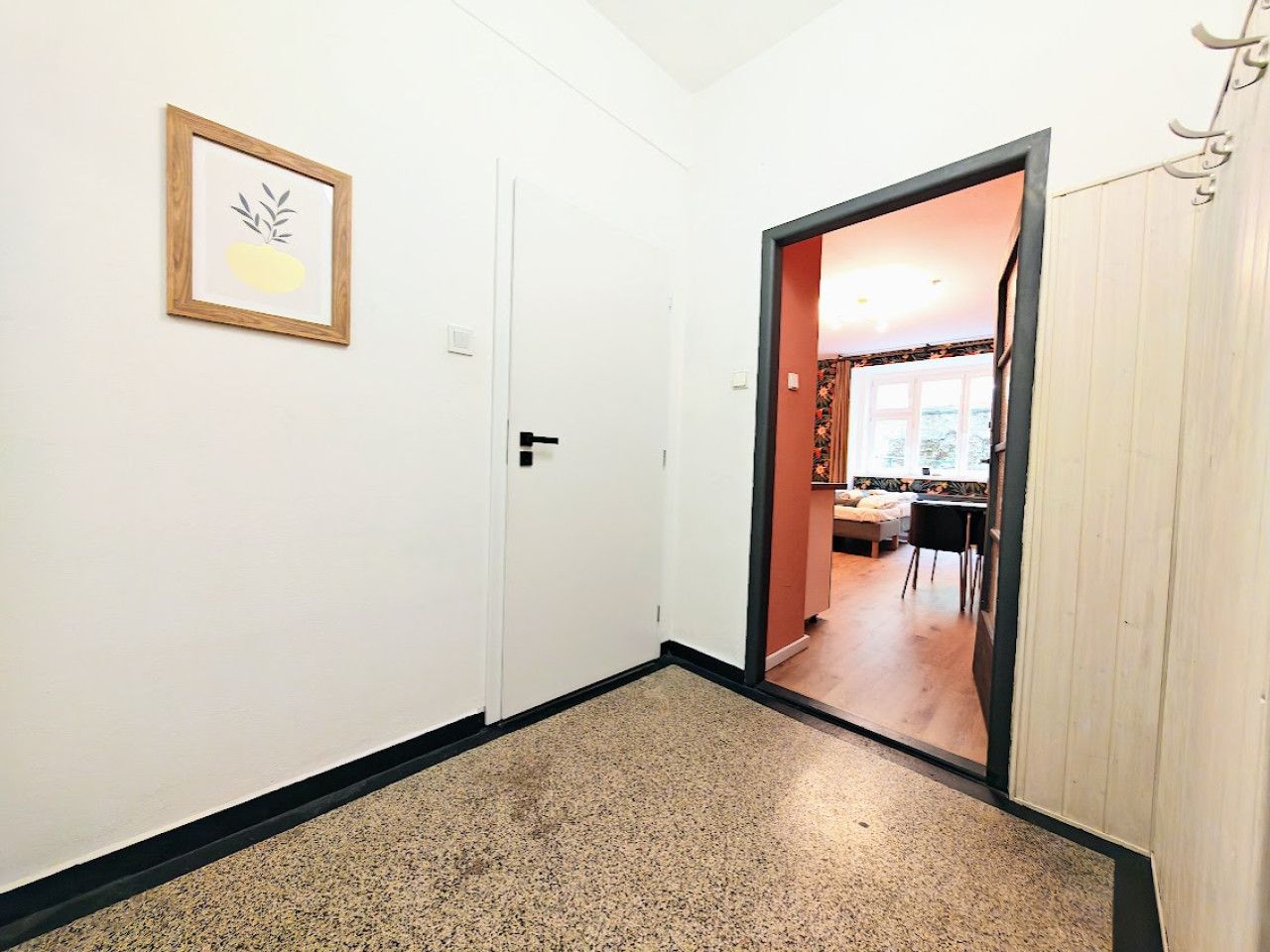 A hallway with a framed picture on the wall, a door leading to a room with a bed and a table, and a wooden door on the right.