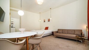 Un espacio habitable compacto con una mesa de comedor, dos sillas, un sofá, una mesita auxiliar y una cama con un cojín rojo.