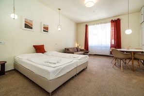 A hotel room with a double bed, a sofa, a dining table with chairs, and a large window with curtains.
