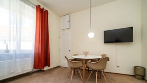 Una habitación pequeña y bien iluminada, con una mesa redonda y cuatro sillas, un televisor montado en la pared, y una puerta con marco blanco.