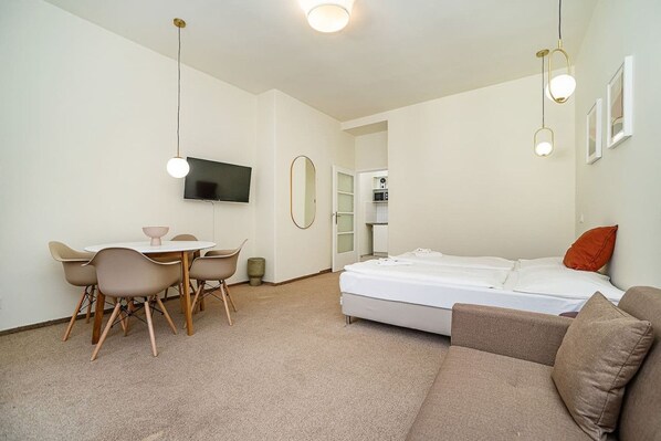 A modern room with a bed, a sofa, a dining table with chairs, a TV mounted on the wall, and a kitchen area visible through an open door.