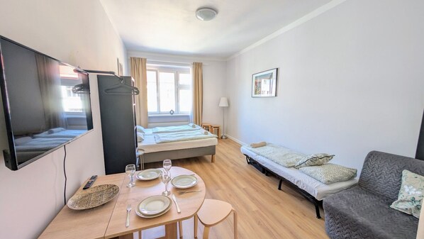 A compact hotel room with a bed, a sofa, a TV, a dining table with chairs, and a small table with a remote control and glasses.