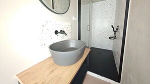 A modern bathroom with a large vessel sink, a wooden countertop, a round mirror, and a shower area with a glass door.