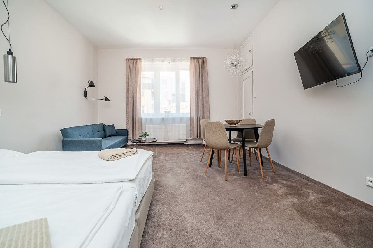 A hotel room with a bed, a blue sofa, a dining table with chairs, a TV mounted on the wall, and a window with curtains.
