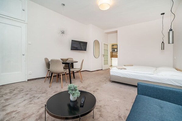 A hotel room with a bed, a blue sofa, a round table with a remote and a plant, a TV on the wall, and a door leading to a kitchen.