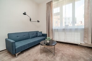 A room with a blue sofa, a round coffee table, a wall-mounted lamp, and a large window with curtains.