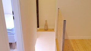Un couloir étroit au plancher en bois, une porte blanche au fond et une petite étagère sur laquelle se trouve un vase.