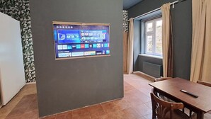 A room with a flat-screen TV mounted on the wall displaying a streaming service interface. There is a window with curtains, a dining table with chairs, and a radiator below the window. - Chic+Contemporary Zizkov Studio (Praha 3)