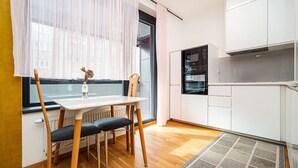 Eine kleine Küche mit weißen Schränken, einem Esstisch mit Stühlen und einem Fenster mit transparenten Vorhängen.