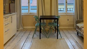 Une pièce compacte comprenant un coin cuisine, deux fenêtres avec rideaux, une petite table avec des chaises et un lit.