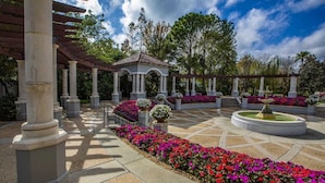 Un espacio al aire libre cubierto con una fuente central, rodeado de macizos de flores y columnas.