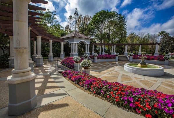 A covered outdoor area with a central fountain, surrounded by flower beds and columns.