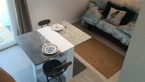 A dining area with a table set for two, a chair, a small kitchen area, and a sofa with decorative pillows.
